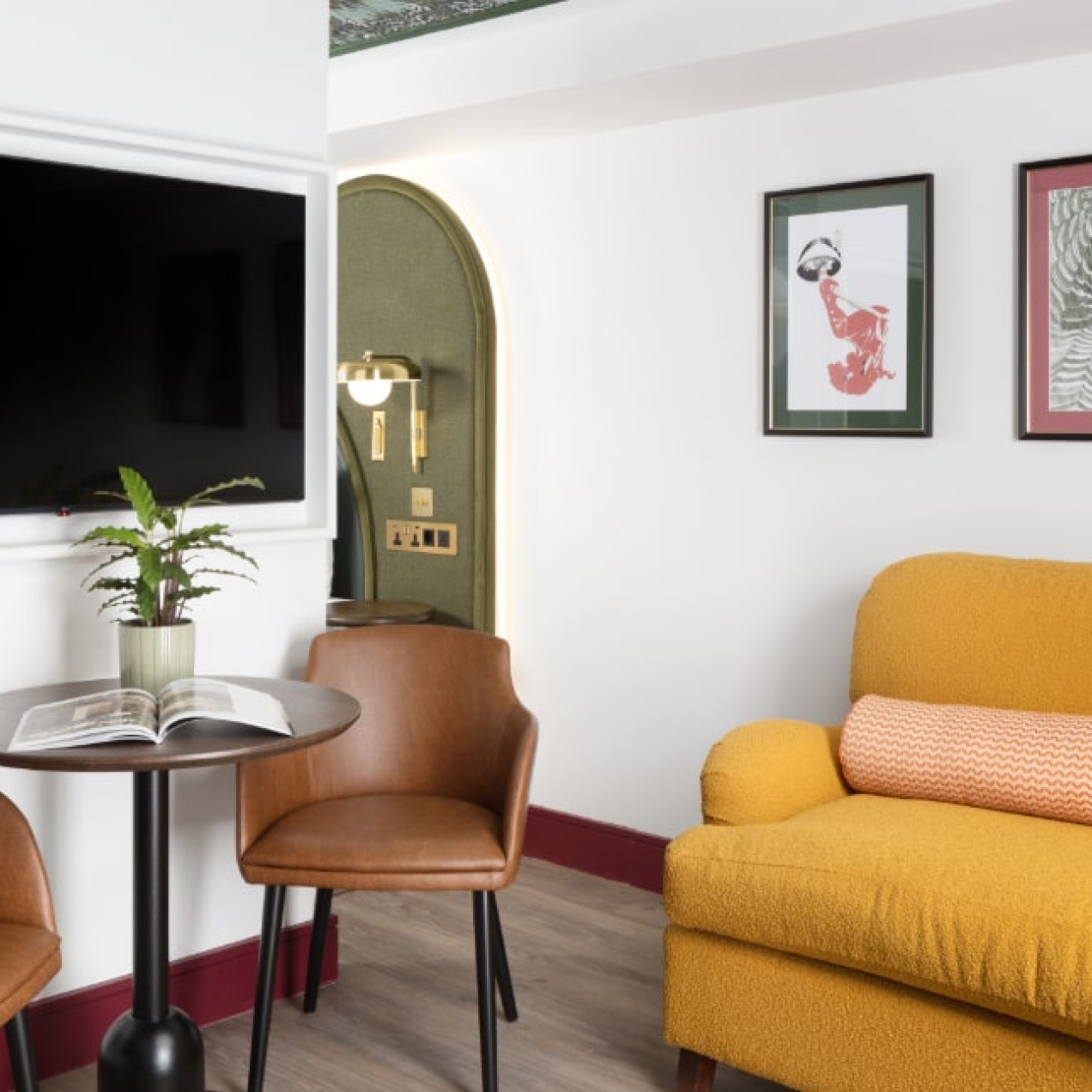 A picture of a hotel suite with white walls with pictures on them and a wall mounted TV, a table and brown chair and a yellow sofa