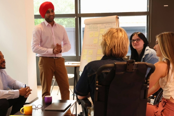 A group of five colleagues: A Black man in a blue shirt sat on a sofa. To his left stands an Asian man with an orange turban and pink shirt stood next to a flip chart. He is smiling at a person with blonde hair who uses a motorised wheelchair. To the