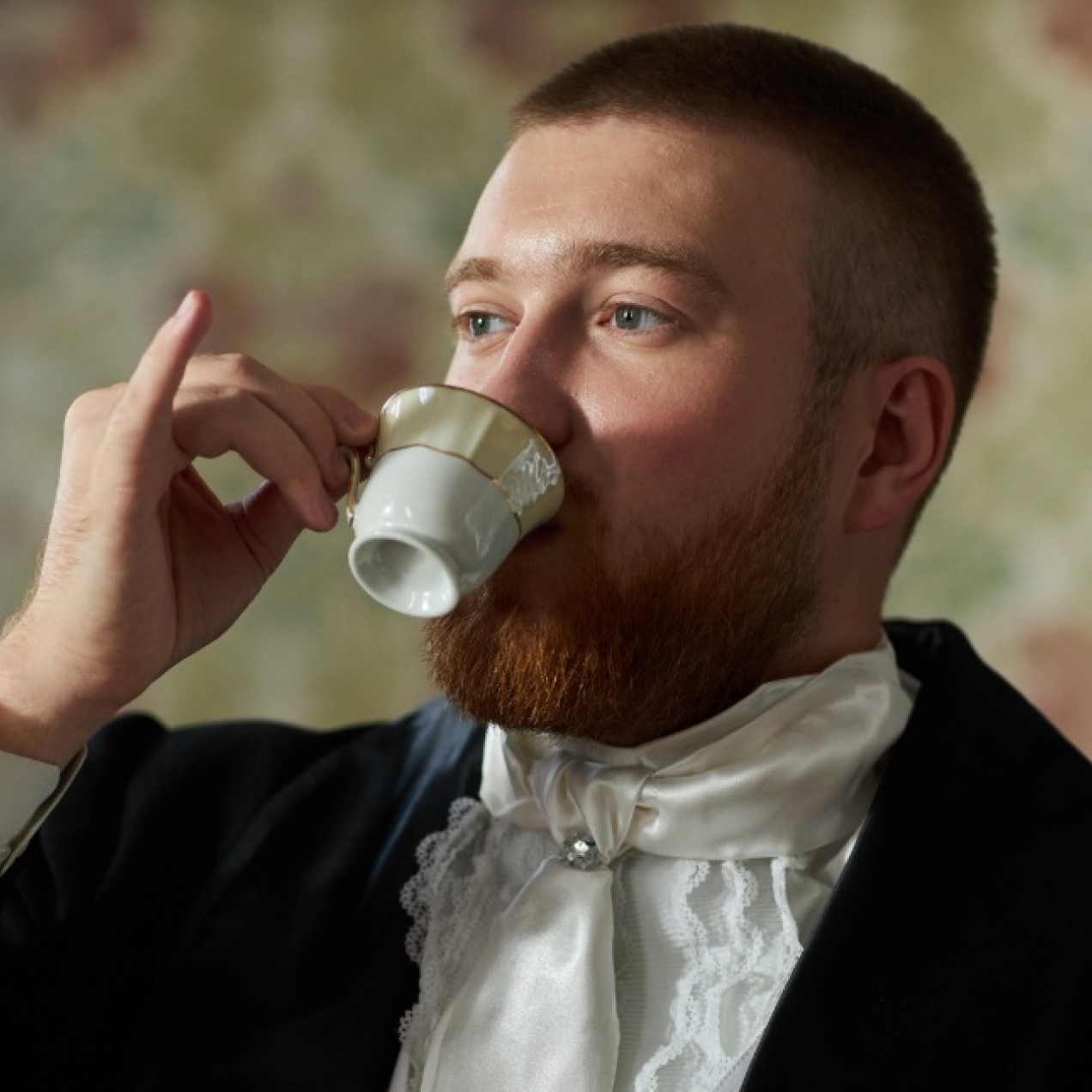 A white man with cropped brown hair and wearing a formal dark jacket and white ruffled shirt holds a porcelain tea cup with his pinkie finger extended.