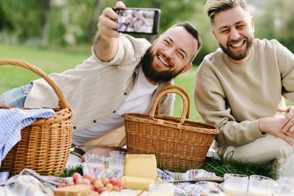 Two white men with beards sit laughing outdoors on a picnic blanket. The man on the left is taking a selfie of them both surrounded by picnic baskets, fruit, bread and drinks.