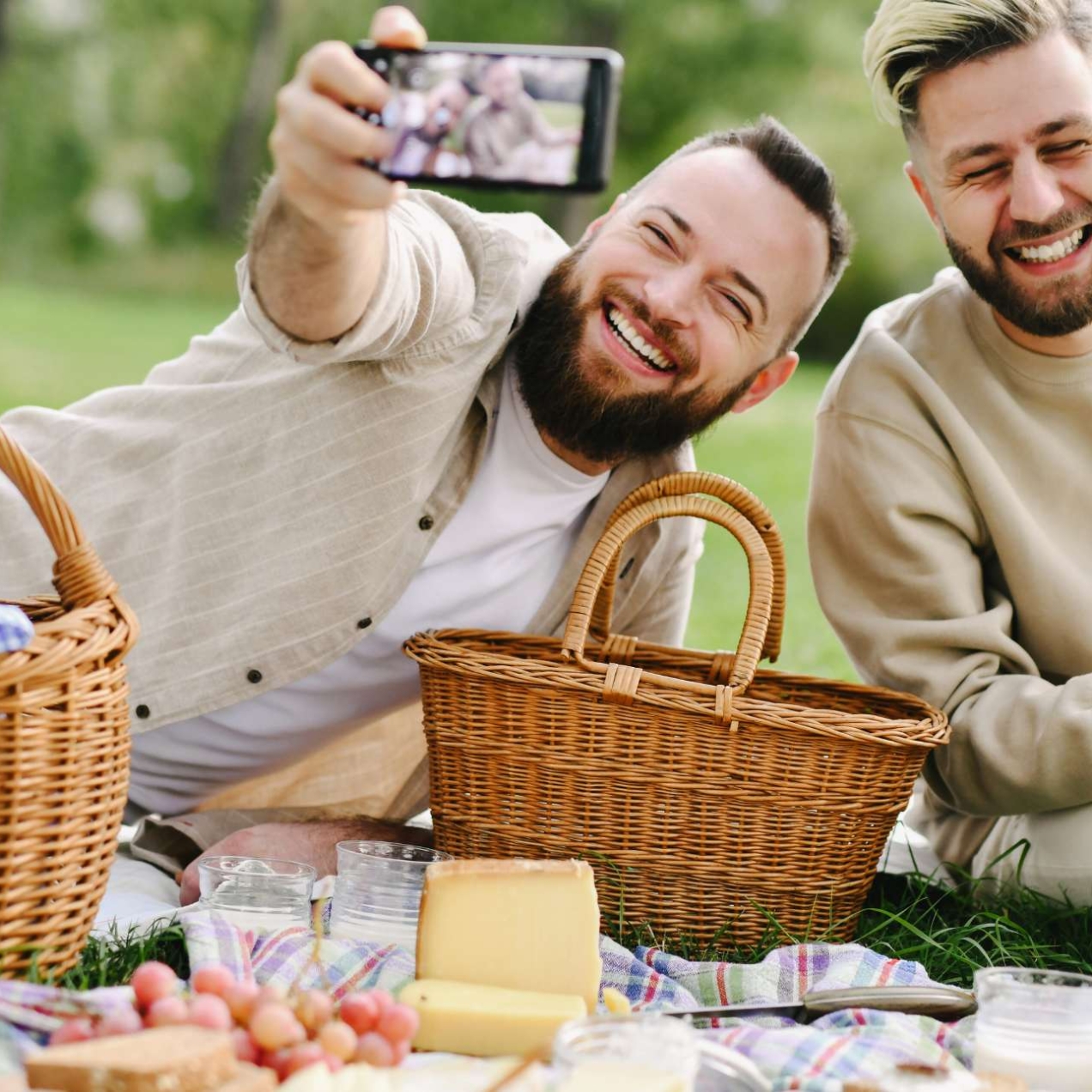 Two white men with beards sit laughing outdoors on a picnic blanket. The man on the left is taking a selfie of them both surrounded by picnic baskets, fruit, bread and drinks.