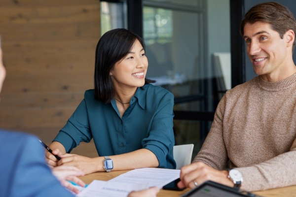 An Asian woman and a white man sit smiling on one side of a board table. Opposite them is the obscured view of another woman