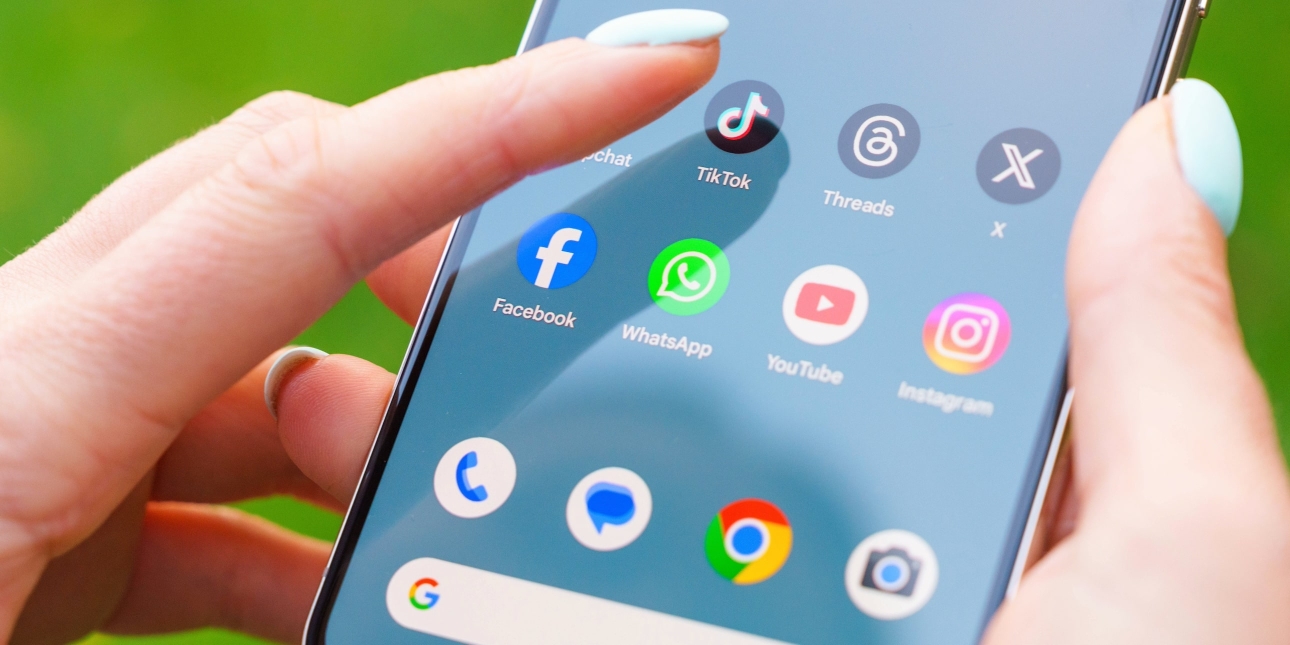 A white finger with eggshell blue nail varnish hovers over the TikTok app on a mobile phone screen. Other icons include Threads, X, Facebook, YouTube and Instagram