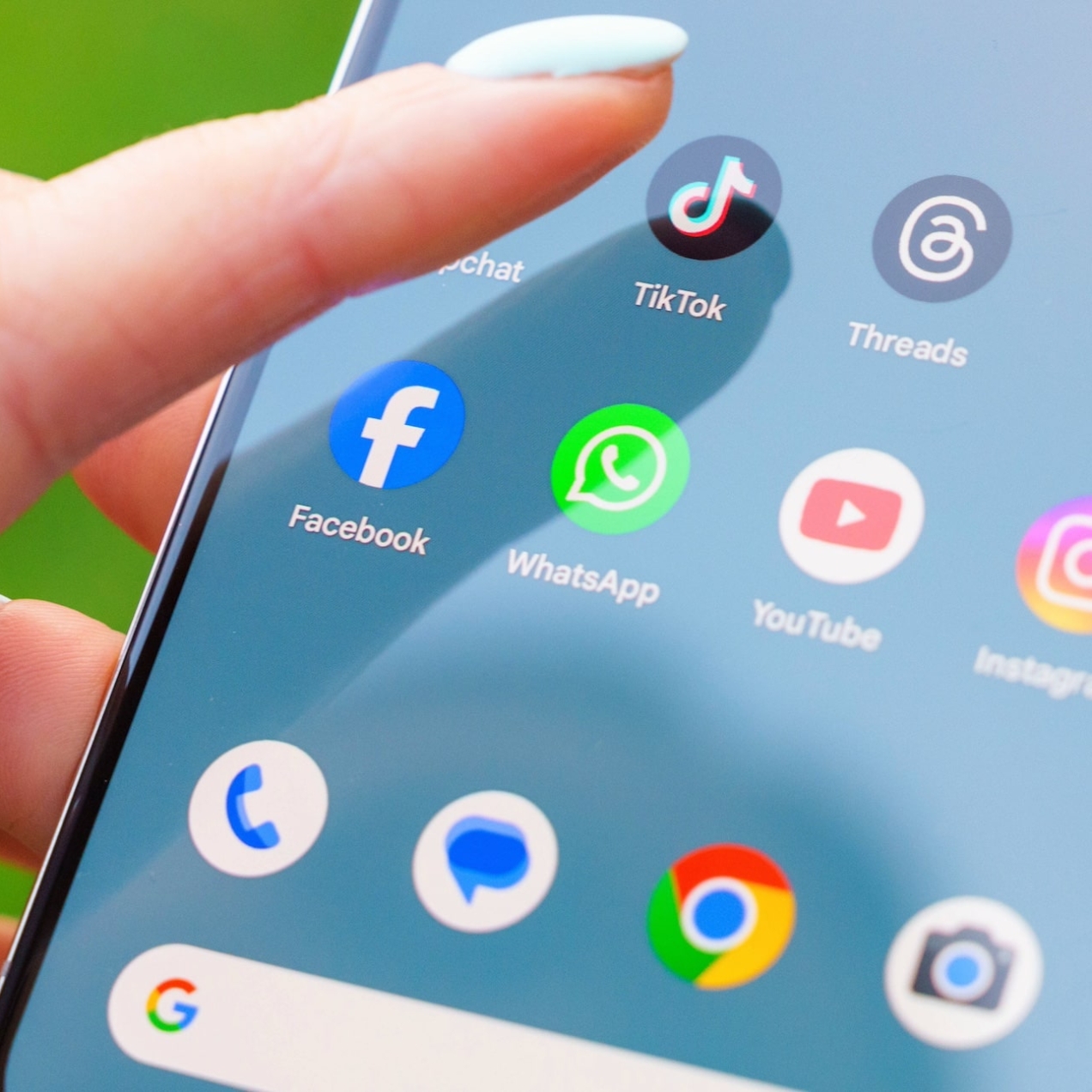 A white finger with eggshell blue nail varnish hovers over the TikTok app on a mobile phone screen. Other icons include Threads, X, Facebook, YouTube and Instagram