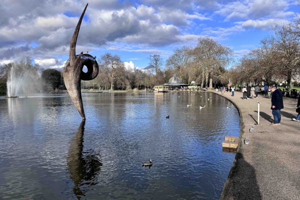 The boating at Victoria Park. White and grey clouds in the sky are reflected into the lake, which is edged with a tree-lined concrete path. People are walking past. A sculpture emerges from the lake. Its form is like a curvy line with a small loop in