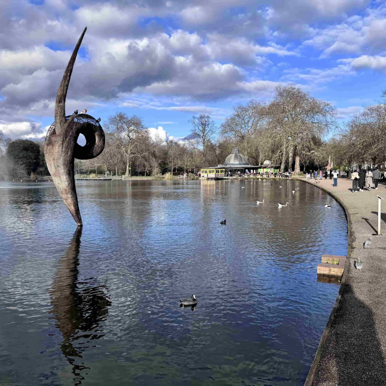 The boating at Victoria Park. White and grey clouds in the sky are reflected into the lake, which is edged with a tree-lined concrete path. People are walking past. A sculpture emerges from the lake. Its form is like a curvy line with a small loop in