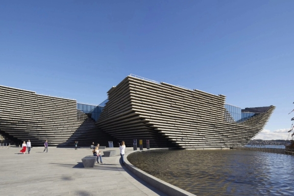 The exterior of the V&A Dundee cladded with lines of pre-cast reconstituted stone panels that run horizontally around curving concrete walls. The shape takes inspiration from cliffs with the prow jutting over the River Tay like a boat. The sky is bri