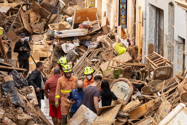 A group of including rescuers walking through a piles of damaged, mud covered furniture and debris on a street