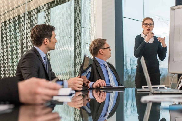 In a modern boardroom a solitary white business woman gives a presentation in front of three white men in suits.