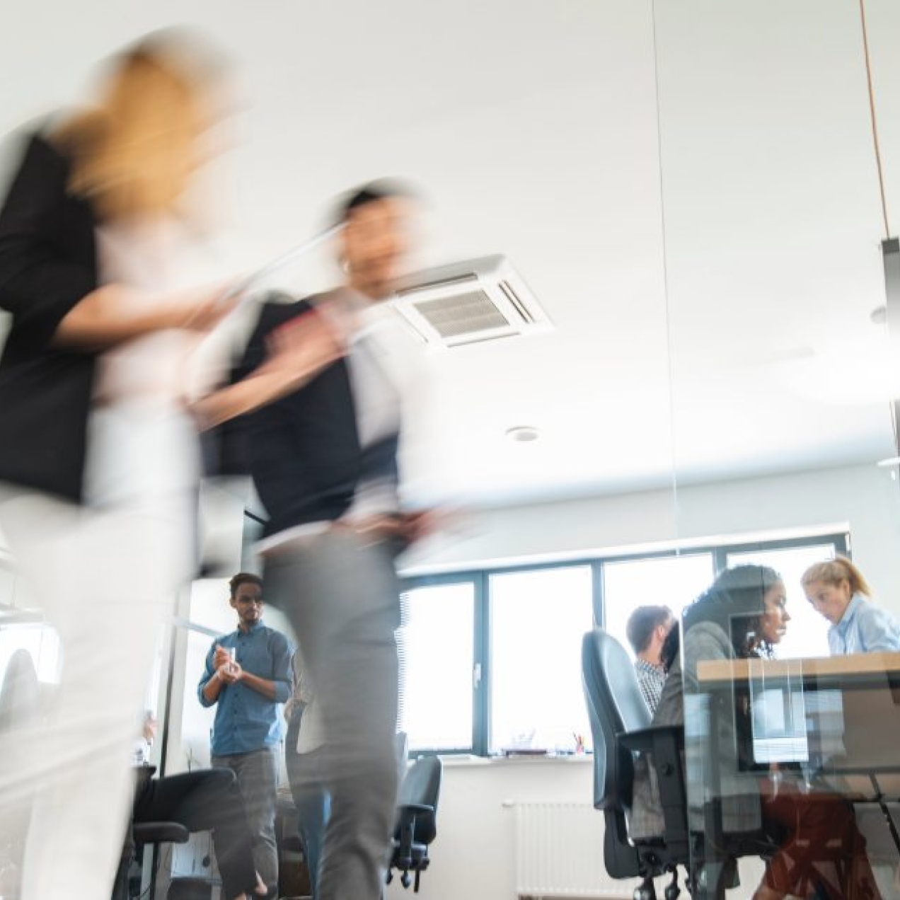 The blurred image of two colleagues walking through an office. Others are sat at desks in the background.