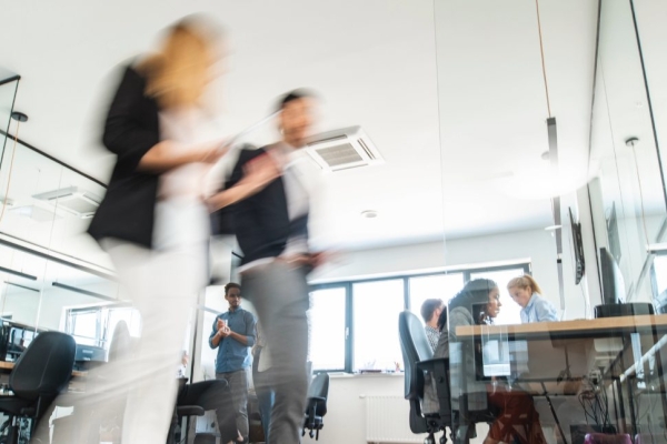 The blurred image of two colleagues walking through an office. Others are sat at desks in the background.