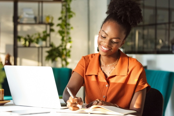 A Black woman wearing an orange blouse smiles while sat at a desk writing in a book with a laptop beside her. She is in a relaxed and trendy office with plants and window.