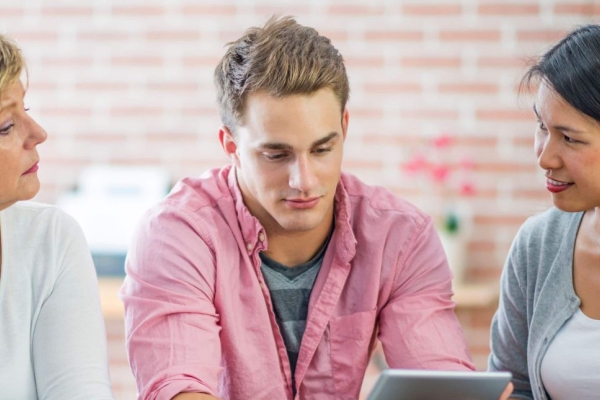 A young white man with short blond hair and pink shirt stares at a tablet. He is flanked on his left by an Asian woman with long dark hair and grey cardigan. On his right is an older white woman with short blonde hair and white top.