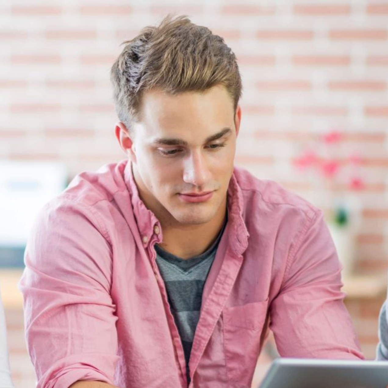 A young white man with short blond hair and pink shirt stares at a tablet. He is flanked on his left by an Asian woman with long dark hair and grey cardigan. On his right is an older white woman with short blonde hair and white top.