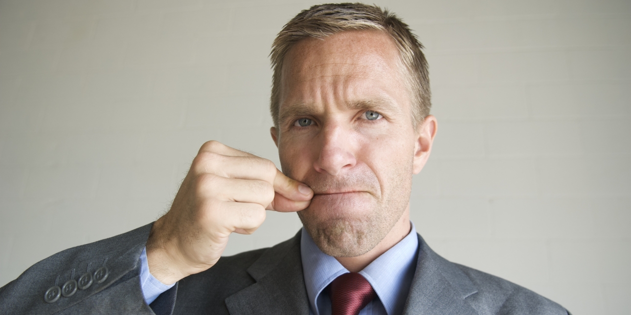 A white businessman with short blond hair and wearing a grey suit with burgundy tie moves his clasped fingers across his mouth as a metaphor for not speaking