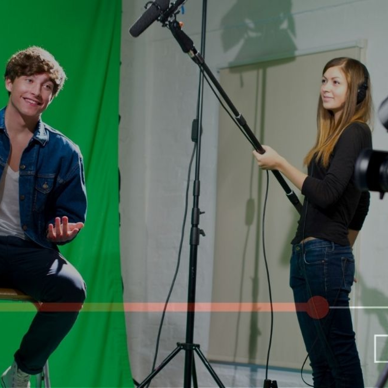 A picture of a young white man sat on a stool in a studio against a green screen. A white woman stood next to him holds a microphone boom while another man crouches behind a camera. The shot is overlaid with video control buttons.