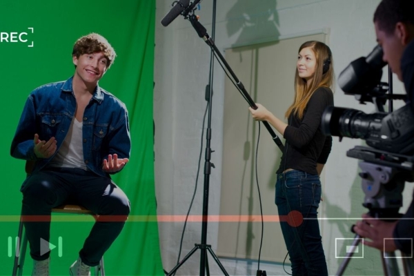 A picture of a young white man sat on a stool in a studio against a green screen. A white woman stood next to him holds a microphone boom while another man crouches behind a camera. The shot is overlaid with video control buttons.