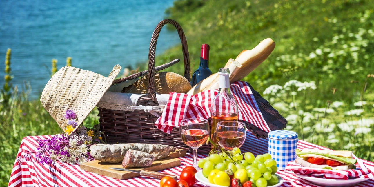 A whicker picnic basket containing red and which check napkins, bread and wine, is sat on a table. The table is also covered in a red and white check table cloth with items of cured meat, glasses of rosé wine, grapes and tomatoes. The background is o