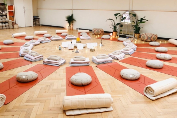Comfy mats and pillows laid out in a circle on the wood floor of a hall. In the centre of the circle are towels, bowls, lamps and other objects.