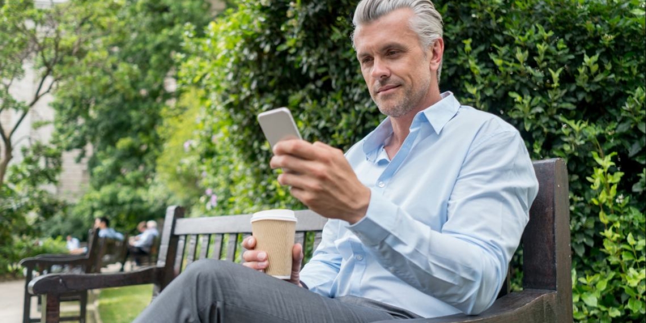 A white man with grey hair, blue shirt and grey trousers sat on a park bench surrounded by trees, looks down at the phone in his left hand. He holds a cup in his right hand.