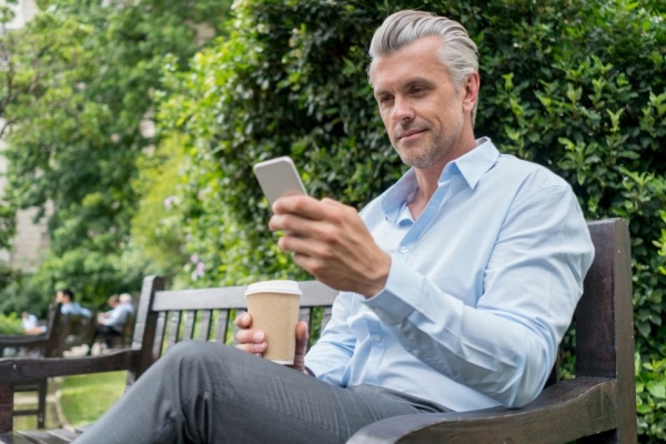 A white man with grey hair, blue shirt and grey trousers sat on a park bench surrounded by trees, looks down at the phone in his left hand. He holds a cup in his right hand.