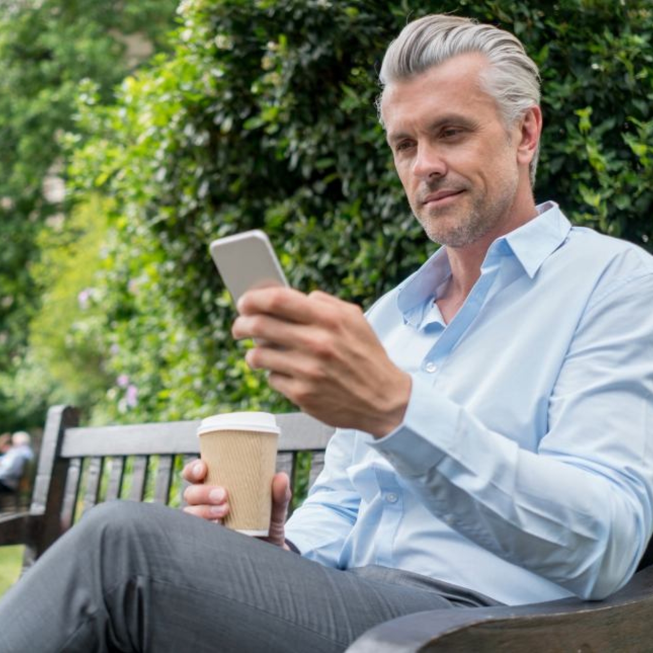 A white man with grey hair, blue shirt and grey trousers sat on a park bench surrounded by trees, looks down at the phone in his left hand. He holds a cup in his right hand.