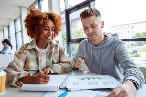 Two college students working together at a desk beside a window. On the left is a Black woman with bronze afro hair and check shirt. On the right is a white man with short brown hair, wearing a grey hoodie.