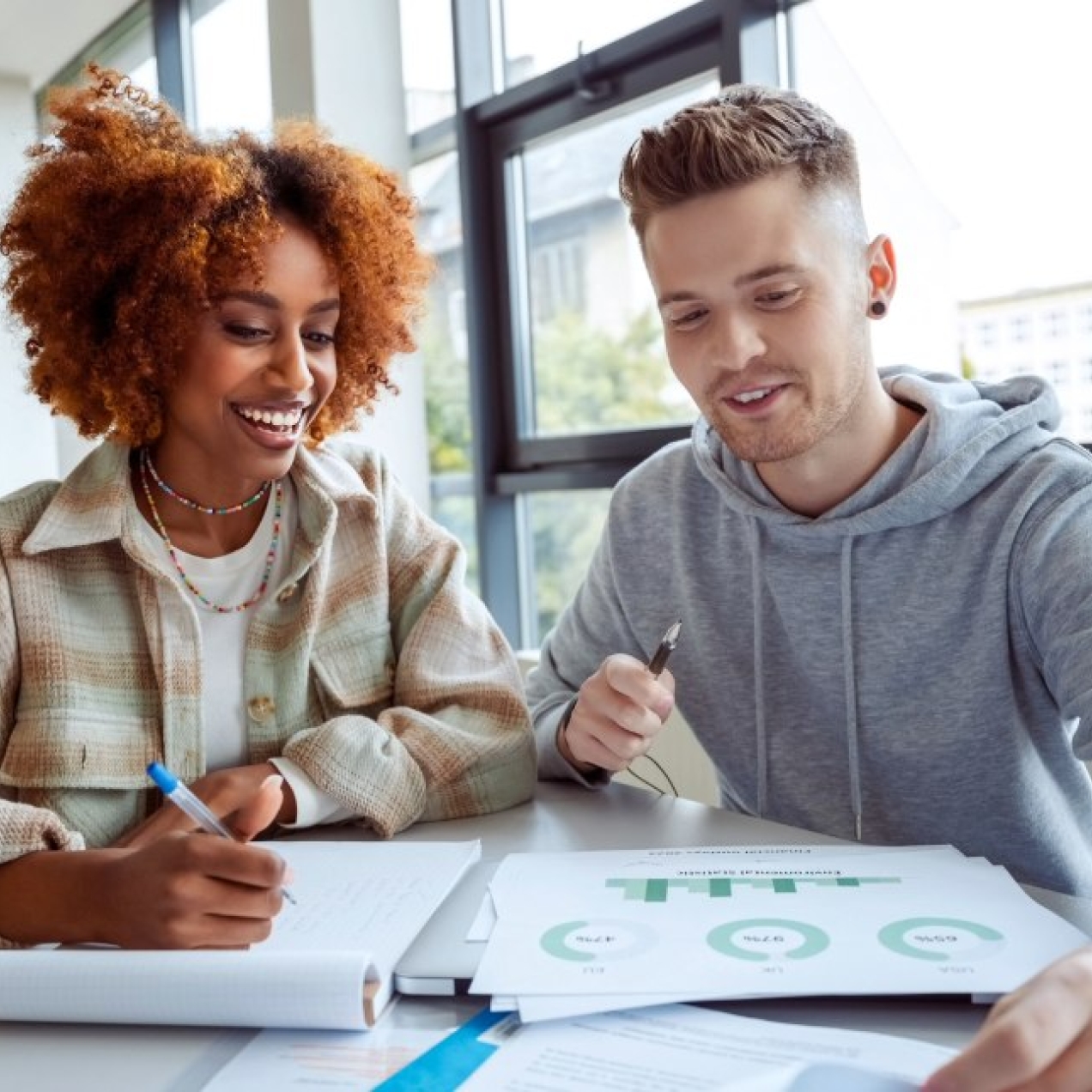 Two college students working together at a desk beside a window. On the left is a Black woman with bronze afro hair and check shirt. On the right is a white man with short brown hair, wearing a grey hoodie.