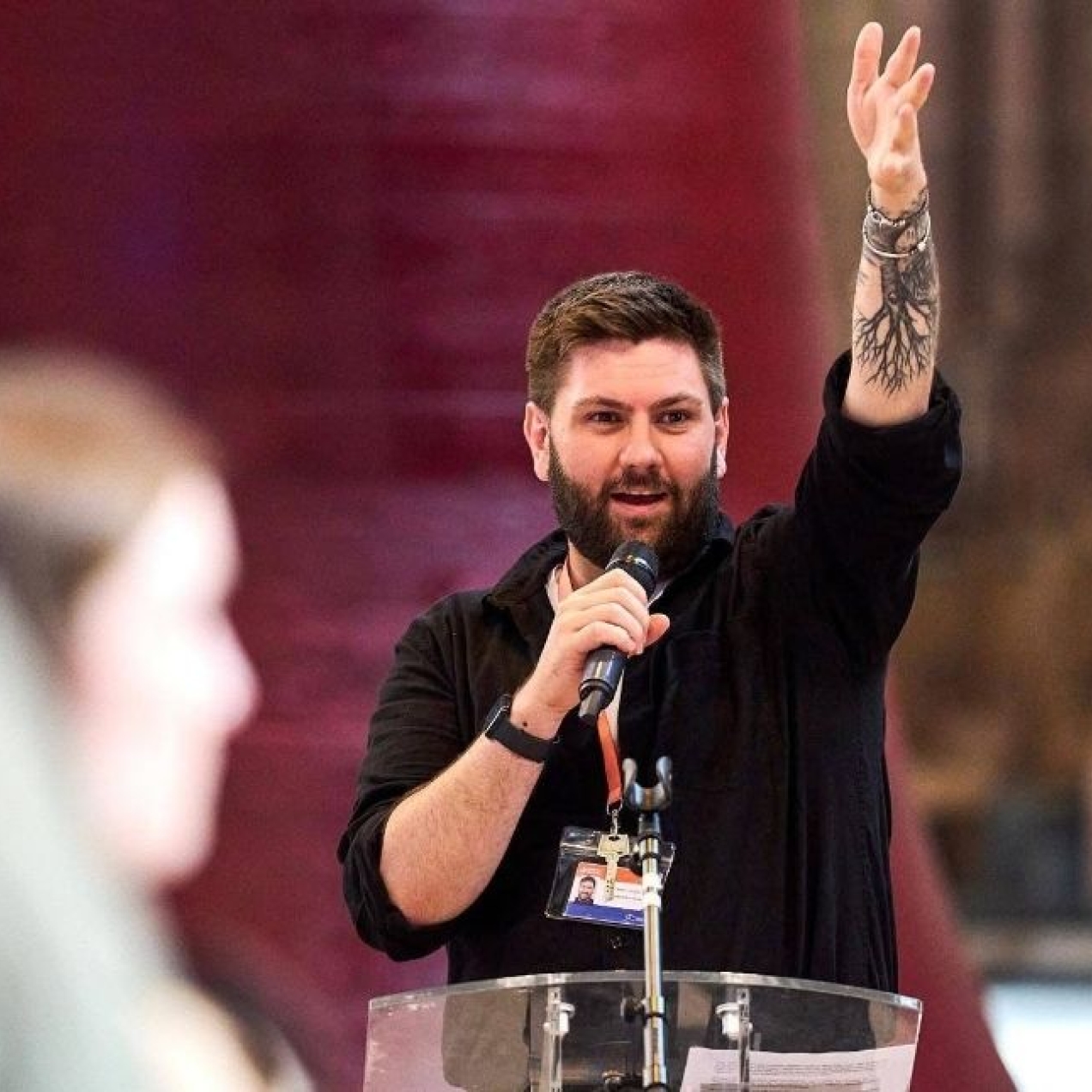 A colour portrait of Daniel Coole in a conference setting holding up his left hand and holding a microphone in her right hand. Daniel is a white man with short dark hair and a tattoo on his arm. Two unidentified people are blurred in the foreground.