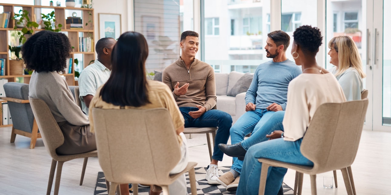 Seven casually dressed people of different ages, genders and ethnicity sit in a circle on chairs. A man on one side of the circle is smiling while talking, the others listen
