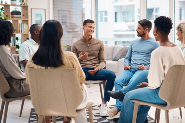 Seven casually dressed people of different ages, genders and ethnicity sit in a circle on chairs. A man on one side of the circle is smiling while talking, the others listen
