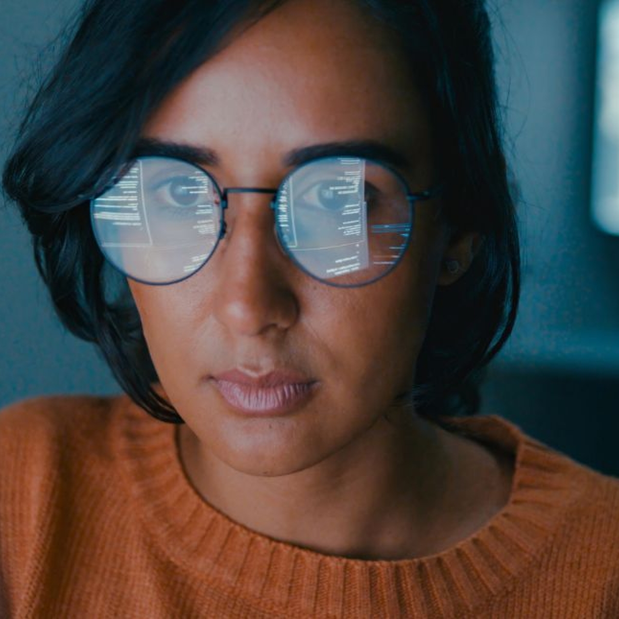 A woman with short dark hair and orange jumper is sat in front of her a computer screen which is reflected in her glasses.