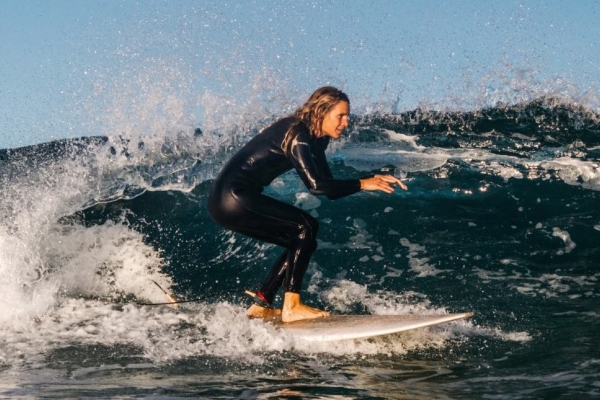 A white female surfer wearing a black all body wetsuit surfing along calm waves with a blue sky behind her