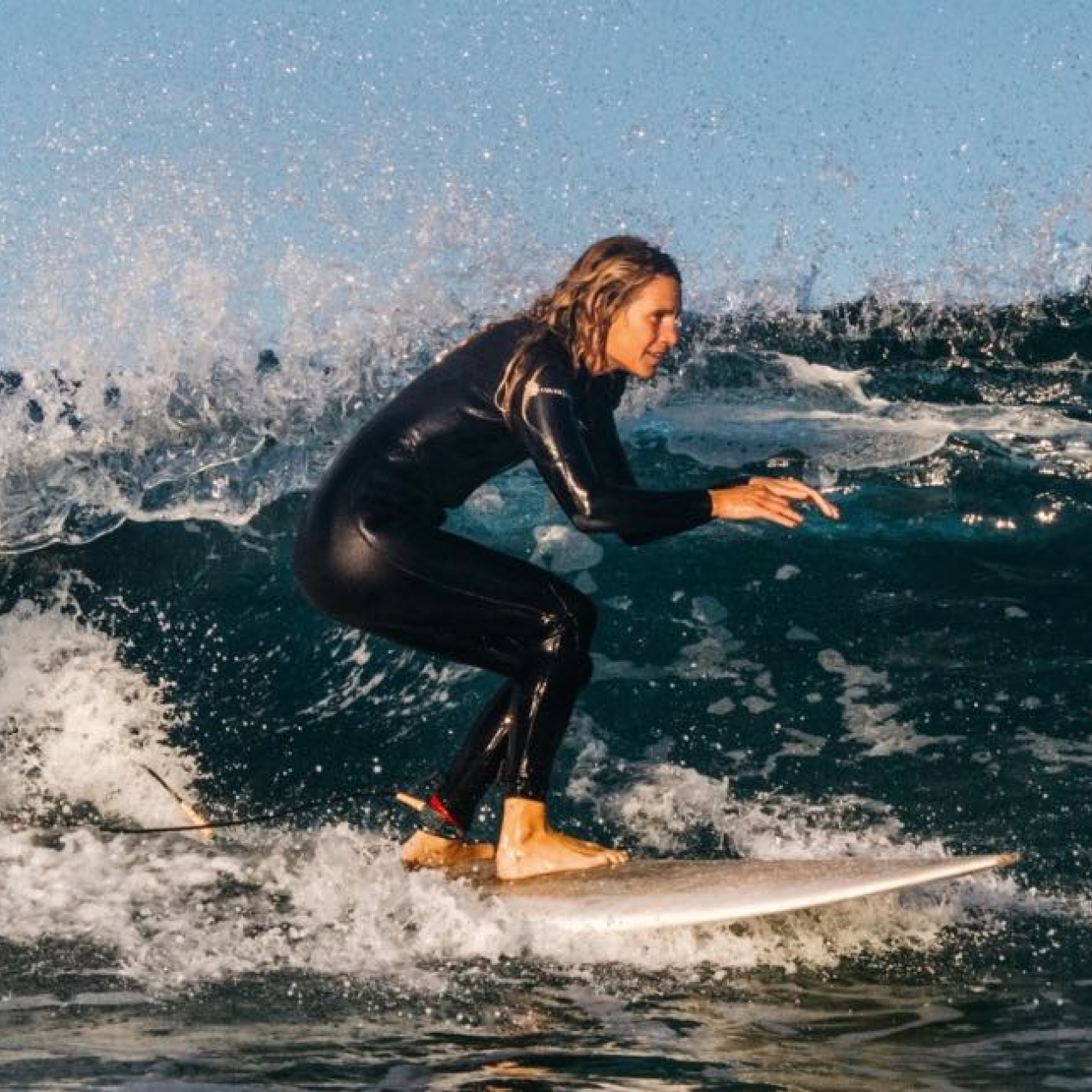 A white female surfer wearing a black all body wetsuit surfing along calm waves with a blue sky behind her