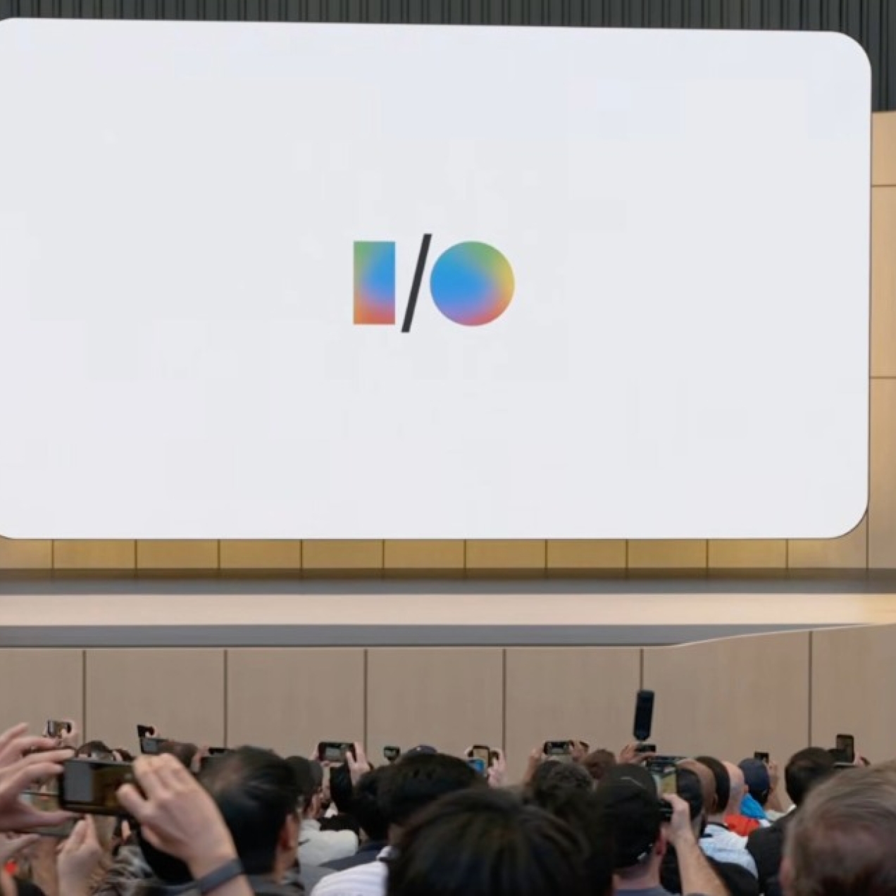 A screenshot from the Google I/O launch event with a shot across the backs of the audience towards a beige stage where a man stands to the right of a screen displaying the I/O logo.