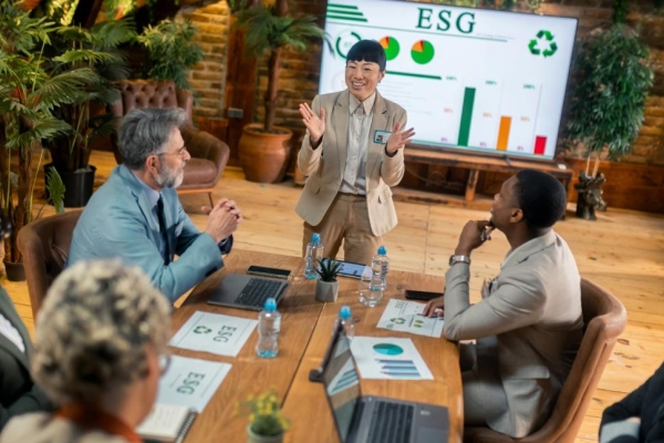 An Asian businesswoman wearing a beige suit is stood in front of a screen featuring the acronym ESG while addressessing a group of male and female colleagues sat at a board table.