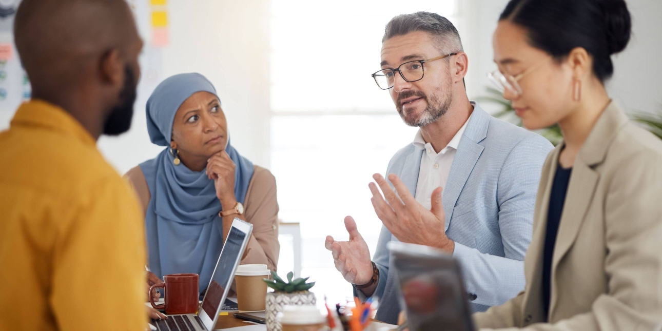In a bright room, four colleagues around a board table. From left to right: a Black man with cropped dark hair, an Asian woman wearing a light blue hijab, a white man with grey hair and an Asian woman with dark hair tied into a bun