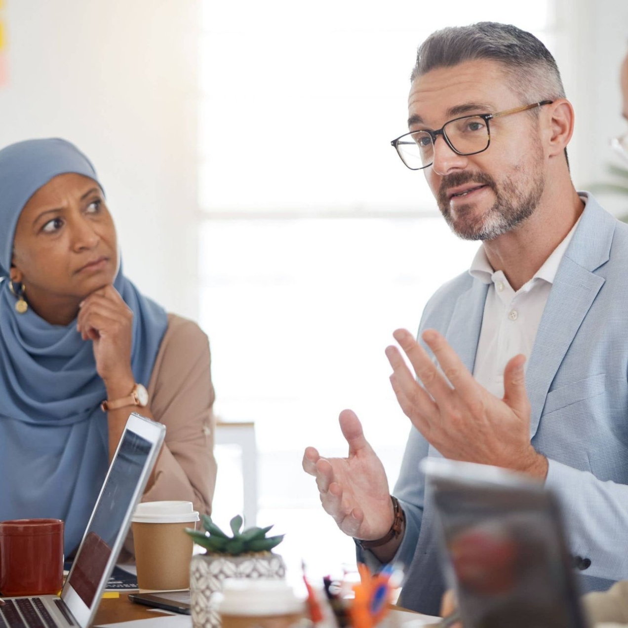 In a bright room, four colleagues around a board table. From left to right: a Black man with cropped dark hair, an Asian woman wearing a light blue hijab, a white man with grey hair and an Asian woman with dark hair tied into a bun