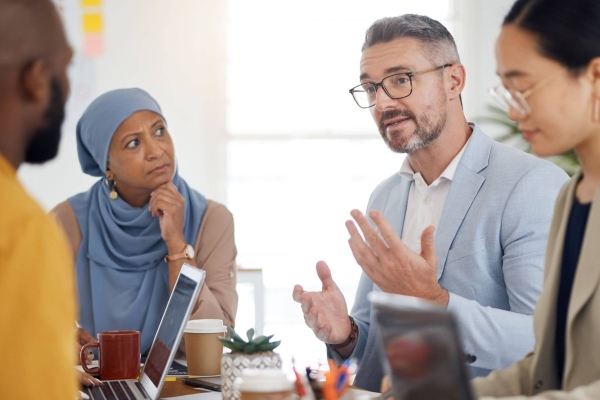 In a bright room, four colleagues around a board table. From left to right: a Black man with cropped dark hair, an Asian woman wearing a light blue hijab, a white man with grey hair and an Asian woman with dark hair tied into a bun