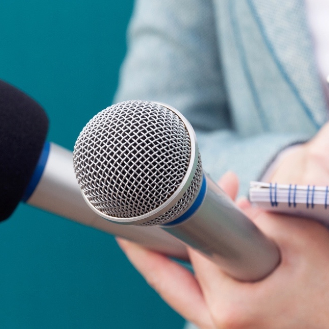 A close up of the torso and white hands of a journalist writing in a notebook while holding two microphones. Her suit is grey, the background is a turquoise