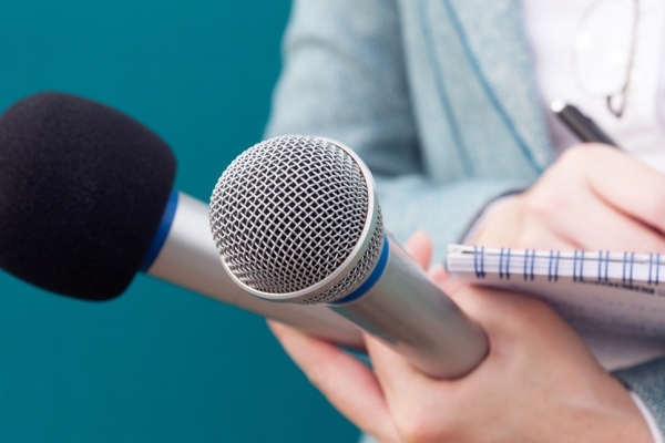 A close up of the torso and white hands of a journalist writing in a notebook while holding two microphones. Her suit is grey, the background is a turquoise