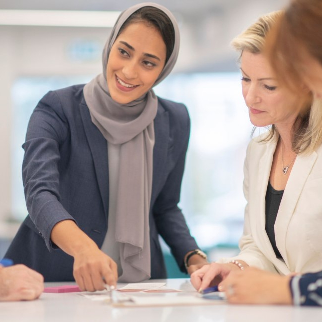 A woman wearing a grey hijab and blue outfit is stood over paperwork on a table while speaking to four male and female colleagues.