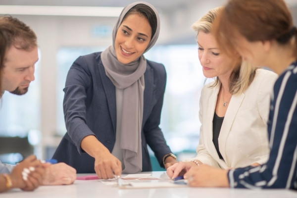 A woman wearing a grey hijab and blue outfit is stood over paperwork on a table while speaking to four male and female colleagues.