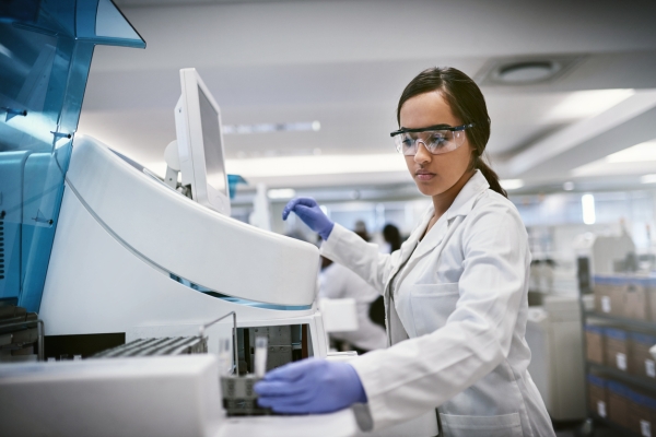 A woman in goggles, lab coat and gloves holds a vial while stood next to a large laboratory computer. The background is blurred but shows a large laboratory space.