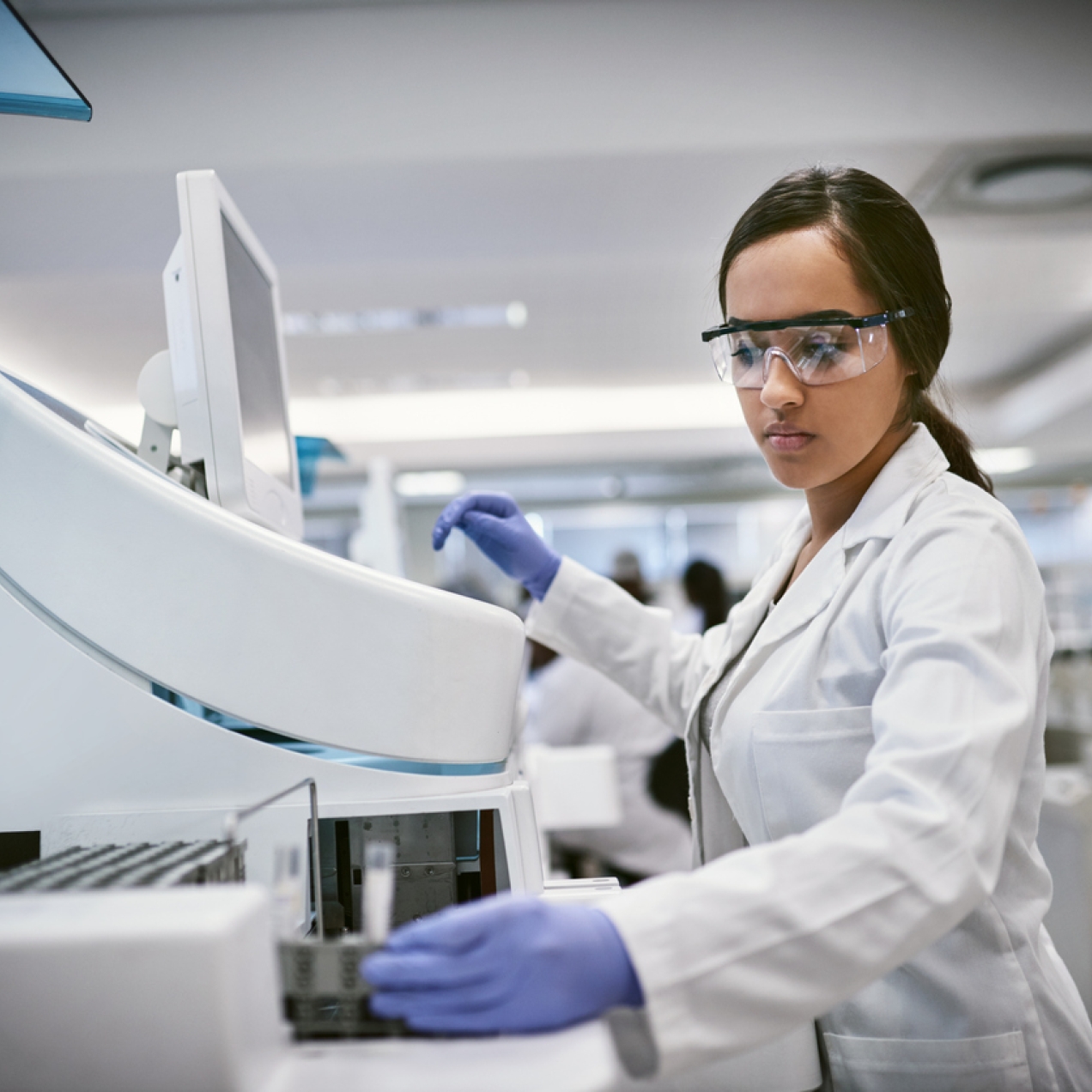 A woman in goggles, lab coat and gloves holds a vial while stood next to a large laboratory computer. The background is blurred but shows a large laboratory space.