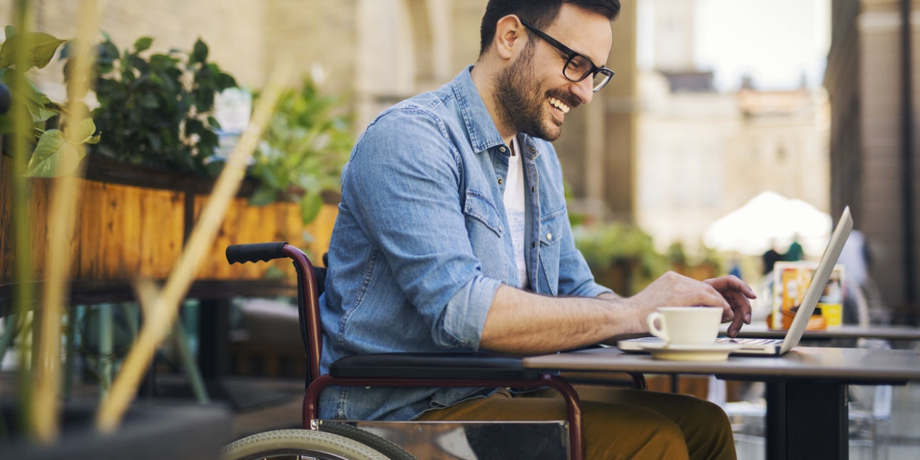 A white man with dark hair, wearing a blue shirt and mustard trousers, is sat in a wheelchair outside a cafe. He is smiling while typing on his laptop on the table.