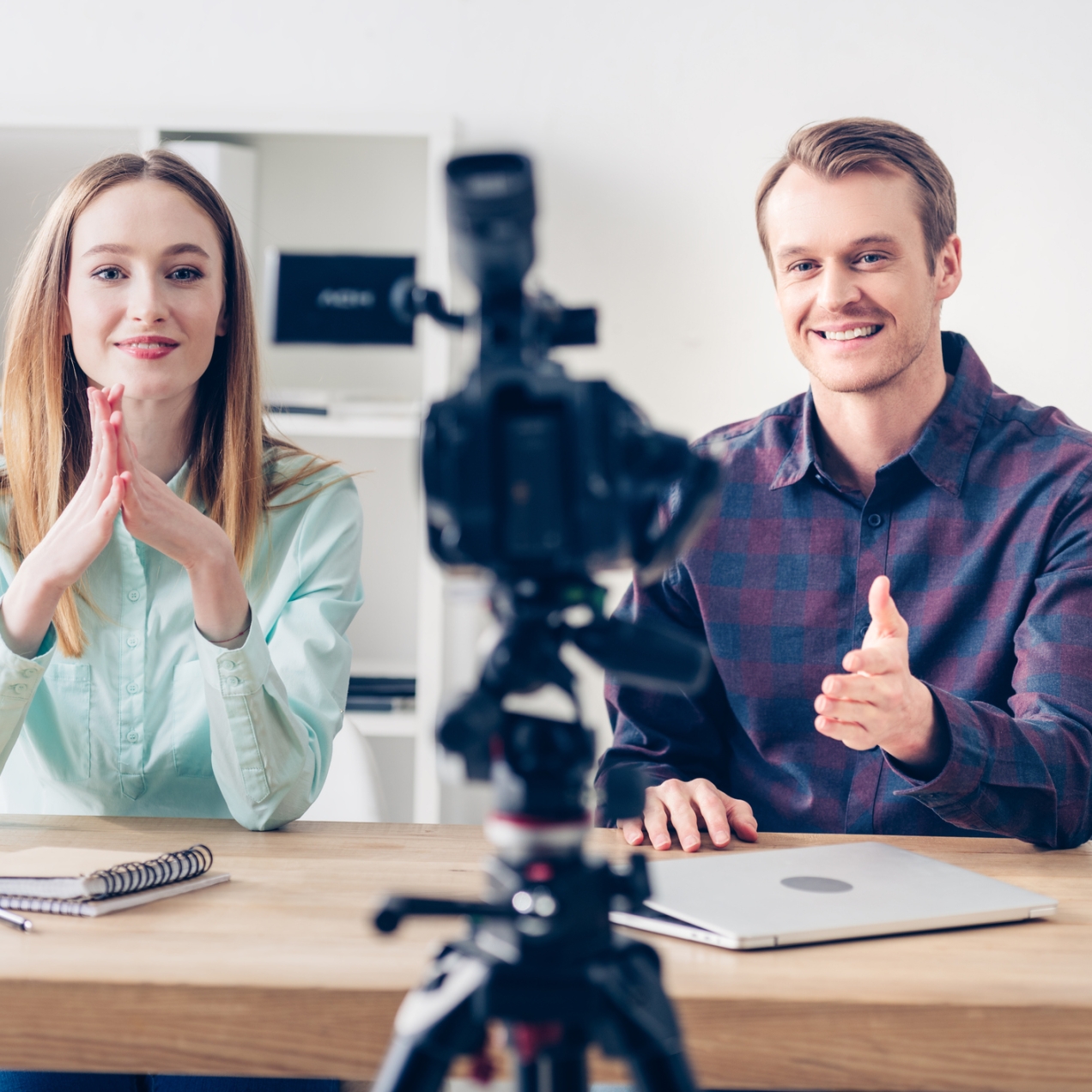 Two business colleagues looking at a video camera. The woman on the left is white with blond hair and smiling. She is holding her palms together with fingers facing upwards. The man on the right is also blond and smiling. He gestures to the camera. T