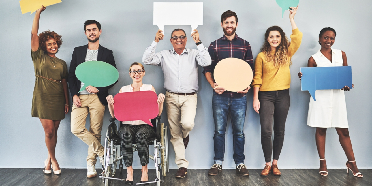 Seven people holding large pieces of card shaped like speech bubbles. Six of the group are standing, one is sat in a wheelchair. The group includes people of different ages, genders and race.