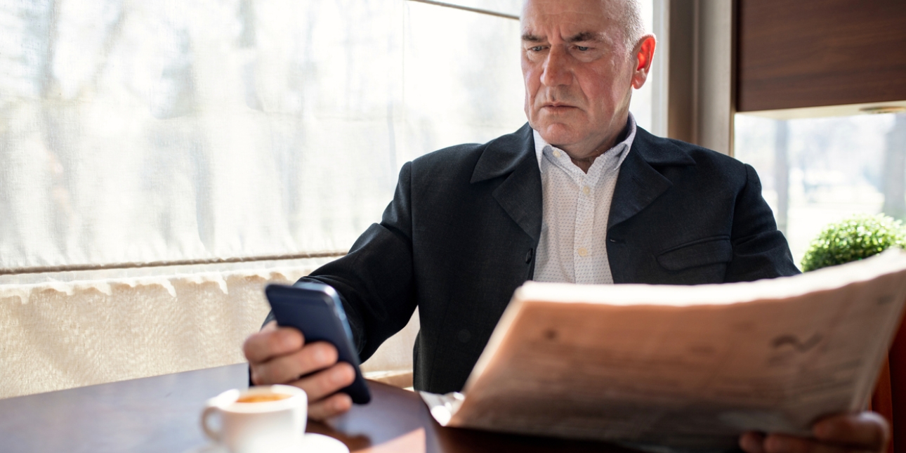 An older, bald white man wearing a smart suit, looks angrily at the mobile phone he is holding in his right hand. In his left hand is a newspaper. An espresso is on the table in front of where he is sat.