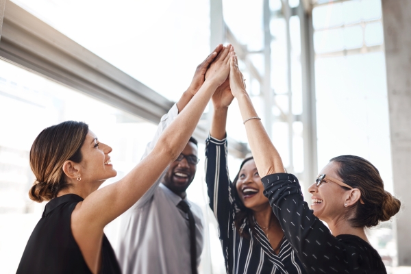 Five business colleagues high-five each other with an up-stretched hand. They comprise of two white women, two black women and one black man. All are dressed in smart-casual clothing.